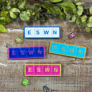 May include: Three rectangular game tiles featuring the letters E, S, W, and N in various color combinations: white with blue, blue with pink, orange with blue, and pink with yellow. Two small translucent dice are positioned near the tiles.