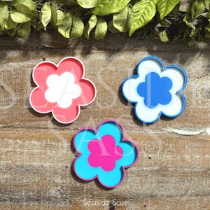 K&ouml;nnte beinhalten: Drei bl&uuml;tenf&ouml;rmige Dekorationen in Rosa, Blau und T&uuml;rkis. Jede Blume hat eine kontrastierende Mitte. Die rosa Blume hat eine wei&szlig;e Mitte, die blaue eine wei&szlig;e Mitte und die t&uuml;rkisfarbene eine rosa Mitte. Auf einer Holzoberfl&auml;che.