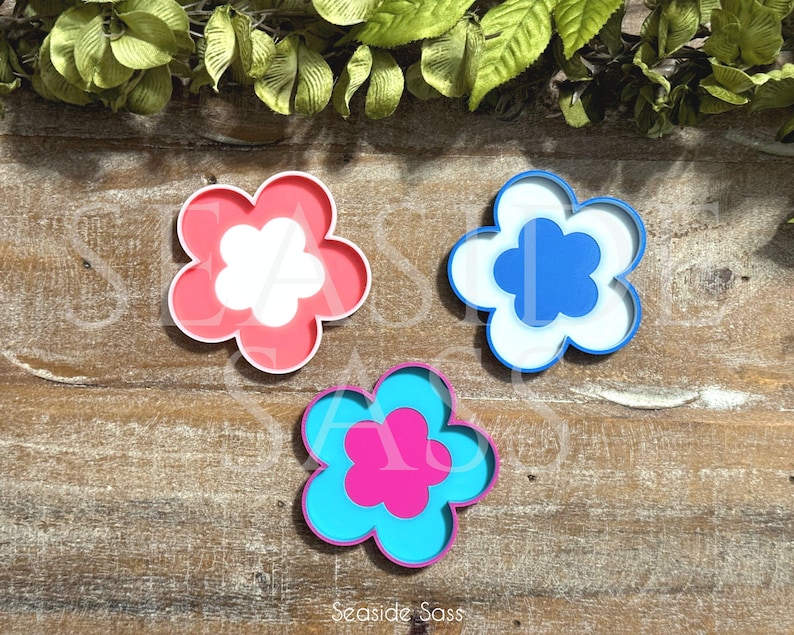 K&ouml;nnte beinhalten: Drei bl&uuml;tenf&ouml;rmige Dekorationen in Rosa, Blau und T&uuml;rkis. Jede Blume hat eine kontrastierende Mitte. Die rosa Blume hat eine wei&szlig;e Mitte, die blaue eine wei&szlig;e Mitte und die t&uuml;rkisfarbene eine rosa Mitte. Auf einer Holzoberfl&auml;che.