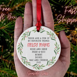 May include: A white ceramic Christmas ornament with a red ribbon, held in a hand. The ornament features a green and red wreath design around the edge. Text includes "THESE ARE A FEW OF MY FAVORITE THINGS" and the name "FINLEY MARIE".