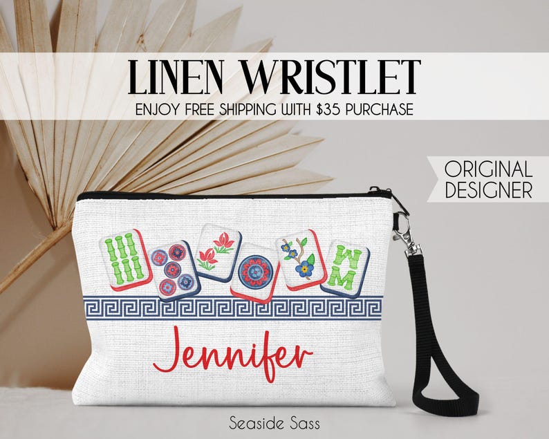 K&ouml;nnte beinhalten: Eine wei&szlig;e Leinen-Handgelenktasche mit einem Grafikdruck von sechs bunten Mahjong-Steinen &uuml;ber einer blauen griechischen Bord&uuml;re. Der Name Jennifer ist in roter Schreibschrift unter der Bord&uuml;re gedruckt. Die Tasche hat eine schwarze Schlaufe und einen Metallverschluss.