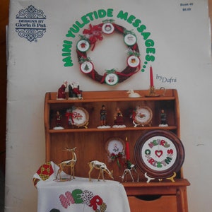 May include: A book titled "Mini Yuletide Messages" by Dafni, featuring a variety of cross-stitch patterns for holiday decorations. The cover shows a wooden shelf with a wreath, ornaments, and a white tablecloth with a cross-stitch design that says "Merry Christmas".
