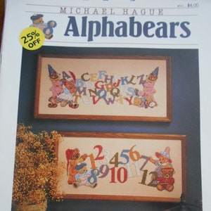 May include: A framed cross-stitch pattern featuring the word "Alphabears" at the top. The image shows two framed cross-stitch samplers. One displays the alphabet with teddy bears, and the other displays numbers with teddy bears.