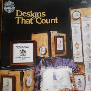 May include: A book titled "Designs That Count" with a collection of cross-stitch patterns featuring various designs, including a frog, a clock, a bird, and a graduation cap. The book cover features a variety of framed cross-stitch projects.