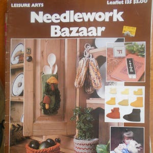 May include: A magazine cover titled "Needlework Bazaar" with a brown background and the text "25 Projects". The cover features a variety of crocheted items, including a basket of avocados, a pot with a plant, a spoon holder, and a pair of oven mitts.