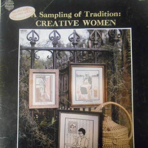 May include: A book cover with a black background and a title that reads "A Sampling of Tradition: CREATIVE WOMEN". The cover features three framed cross-stitch designs and a woven basket.