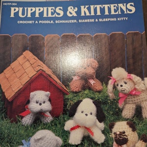 May include: A crochet pattern book featuring a variety of adorable crocheted puppies and kittens. The book cover features a red doghouse with a brown roof, a white poodle, a black and white schnauzer, a Siamese cat, and a sleeping kitten.