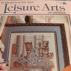 May include: A framed cross-stitch depicting a cozy porch scene with two rocking chairs, flowers, and a table. The magazine cover reads "Leisure Arts" and "31 Projects in this Issue!" The image is framed with a brown border and a blue mat. A violin is in the foreground.
