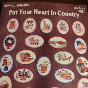 May include: A book cover with the title "Put Your Heart In Country" and various oval-shaped cross-stitch designs. The designs feature country-themed images like rocking chairs, flowers, and farm scenes.