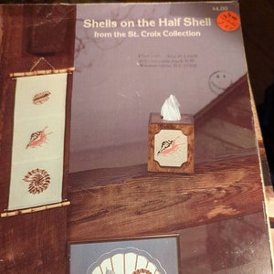 May include: A collection of seashell-themed crafts, including a framed seashell design, a tissue box, and a wall hanging. The wall hanging features three embroidered seashells. The framed design is a seashell fan with various shell illustrations.