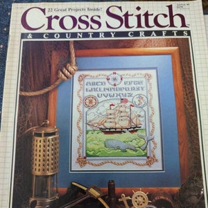 May include: A magazine cover for Cross Stitch & Country Crafts featuring a framed cross stitch design of a ship sailing on a blue sea with a compass and a nautical rope border. The magazine cover also features a title card with the text "Cross Stitch & Country Crafts" and the text "22 Great Projects Inside!"