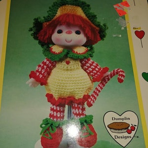 May include: Crochet pattern for a Peppermint Stick doll. The doll is wearing a red and white dress, a green hat, and red shoes. The pattern is from Dumplin Designs.