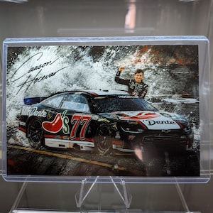 May include: A trading card featuring a black and white race car with the number 77 and a red chili pepper graphic. The car is branded with Dente and Ride Dente logos. A driver stands behind the car with a fist raised. Carson Hocevar is signed.