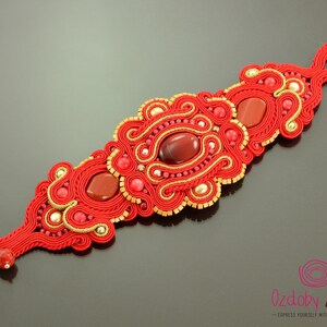 May include: Red and gold bracelet with a red gemstone and intricate woven design. "Ozdoby Ziemi" is written on the bottom of the image.