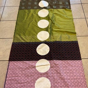 Può includere: Un pannello di tessuto con un motivo ripetitivo di cerchi bianchi su sfondo marrone, seguito da un pannello verde pieno, un pannello a pois bianco e nero, un pannello a pois rosa e nero e un pannello rosso pieno. Ogni pannello ha un grande cerchio bianco al centro.