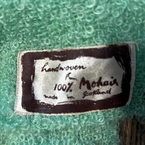 May include: Close-up of a textured, light teal fabric with a sewn-in label. The label reads "Handwoven for 100% Mohair Made in Scotland" in a dark brown frame. The fabric has a soft, looped texture.