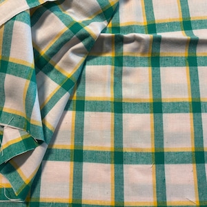 May include: White fabric with a green and yellow plaid pattern. The fabric is folded over, showing the pattern in detail. The plaid design features intersecting green and yellow lines on a white background, creating a grid-like pattern.
