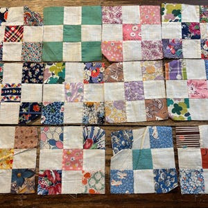 Op de afbeelding: Een verzameling kleine, vierkante patchwork quiltblokken. Elk blok heeft een rasterpatroon van kleurrijke bloemen- en patroonstoffen, afgewisseld met witte vierkanten. De stoffen omvatten verschillende tinten blauw, roze, groen en rood.