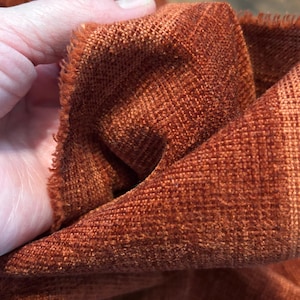 Könnte beinhalten: Nahaufnahme eines strukturierten, rostfarbenen Stoffes. Der Stoff hat ein gewebtes Aussehen mit einer leicht ausgefransten Kante. Die Farbe ist ein warmer, erdiger Ton, der auf ein Textil aus Naturfasern hindeutet.