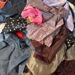 Könnte beinhalten: Ein Haufen bunter Seidenstoffreste in verschiedenen Blau-, Rosa-, Lila-, Braun- und Weißtönen. Einige Teile haben Muster, darunter Blumen und Tupfen. Der Stoff scheint glänzend und glatt zu sein.