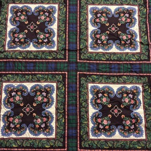 May include: A quilted textile featuring a plaid pattern in green and blue, with four large square floral designs. Each square has a dark center with a floral arrangement, surrounded by a scalloped blue border and green leaves. Pink ribbon and floral accents are on the edges.