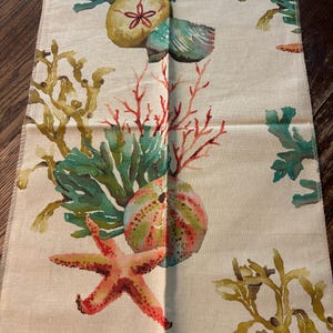 May include: A fabric sample with a sea life pattern. The design features a starfish, sea urchins, and coral in shades of red, green, and yellow on a cream background. The fabric has the text "SEALIFE MULTI" and "Temple Furniture" printed on a tag.