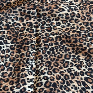 Puede incluir: Primer plano de una tela con estampado de leopardo. El patrón presenta manchas negras irregulares sobre un fondo marrón y marrón. La tela parece suave y flexible, con pliegues y arrugas visibles.