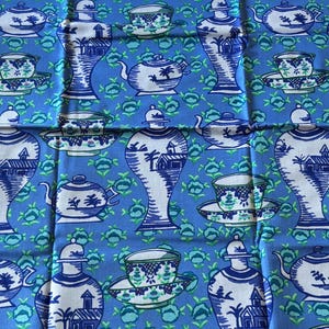 May include: A blue fabric featuring a repeating pattern of white and dark blue teapots, lidded jars, and teacups on saucers. The background is a vibrant blue decorated with small, repeating green floral motifs throughout the entire textile design.