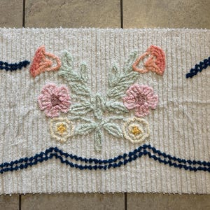 Puede incluir: Alfombra rectangular de color crema con diseño floral. La alfombra presenta un ramo central de flores rosas, melocotón y amarillas con hojas verdes. Ribetes azul oscuro acentúan los bordes superior e inferior. La alfombra está hecha de un material texturizado.