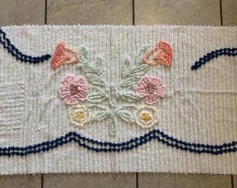 May include: Cream-colored, rectangular rug with a floral design. The rug features a central bouquet of pink, peach, and yellow flowers with green leaves. Dark blue trim accents the top and bottom edges. The rug is made of a textured, looped material.