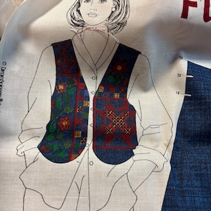 May include: A fabric panel featuring a sketched illustration of a woman wearing a vest. The vest is navy blue with floral and geometric patterns in red, yellow, and green. The woman is wearing a white shirt. The word "FUN" is visible in red.