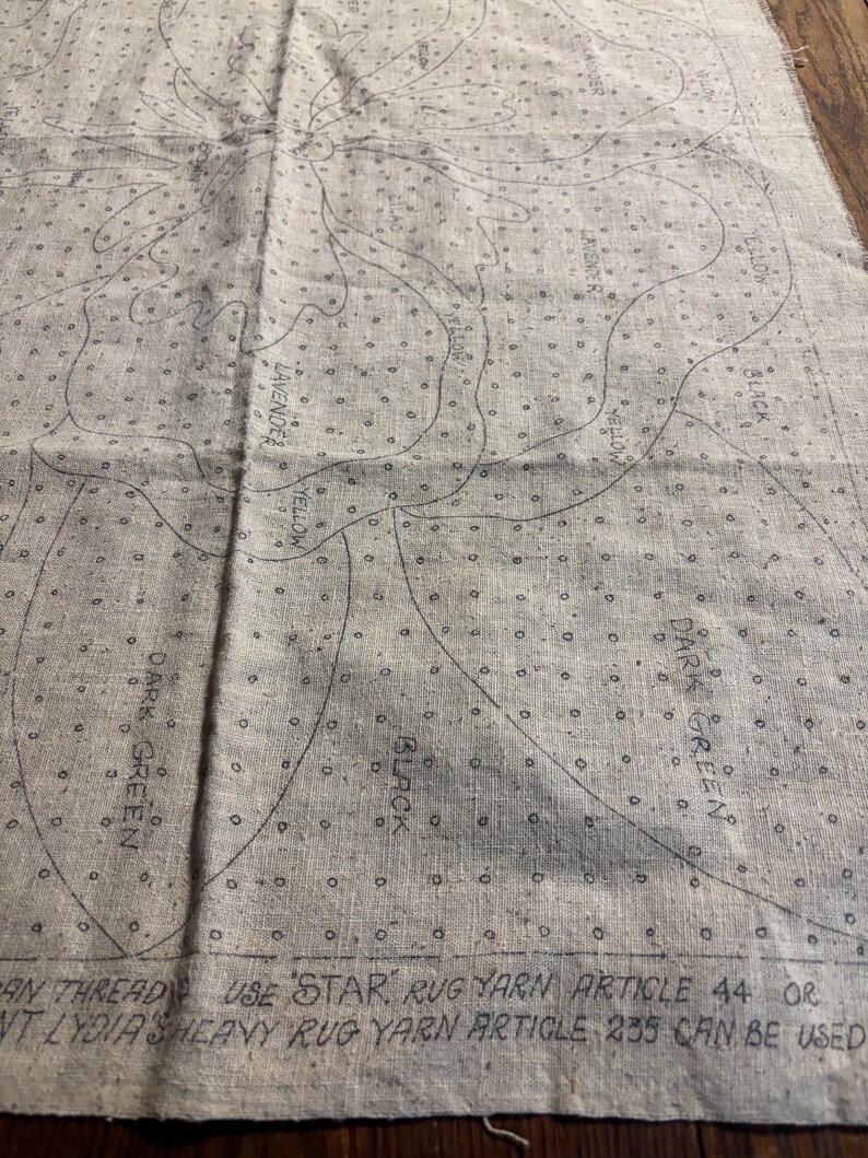 May include: A close-up of a beige fabric with a printed pattern of a flower. The pattern is outlined in black and includes the words "Yellow", "Lavender", "Black", "Dark Green", and "Use 'Star' rug yarn article 44 or 'Lydia's heavy rug yarn article 235 can be used."