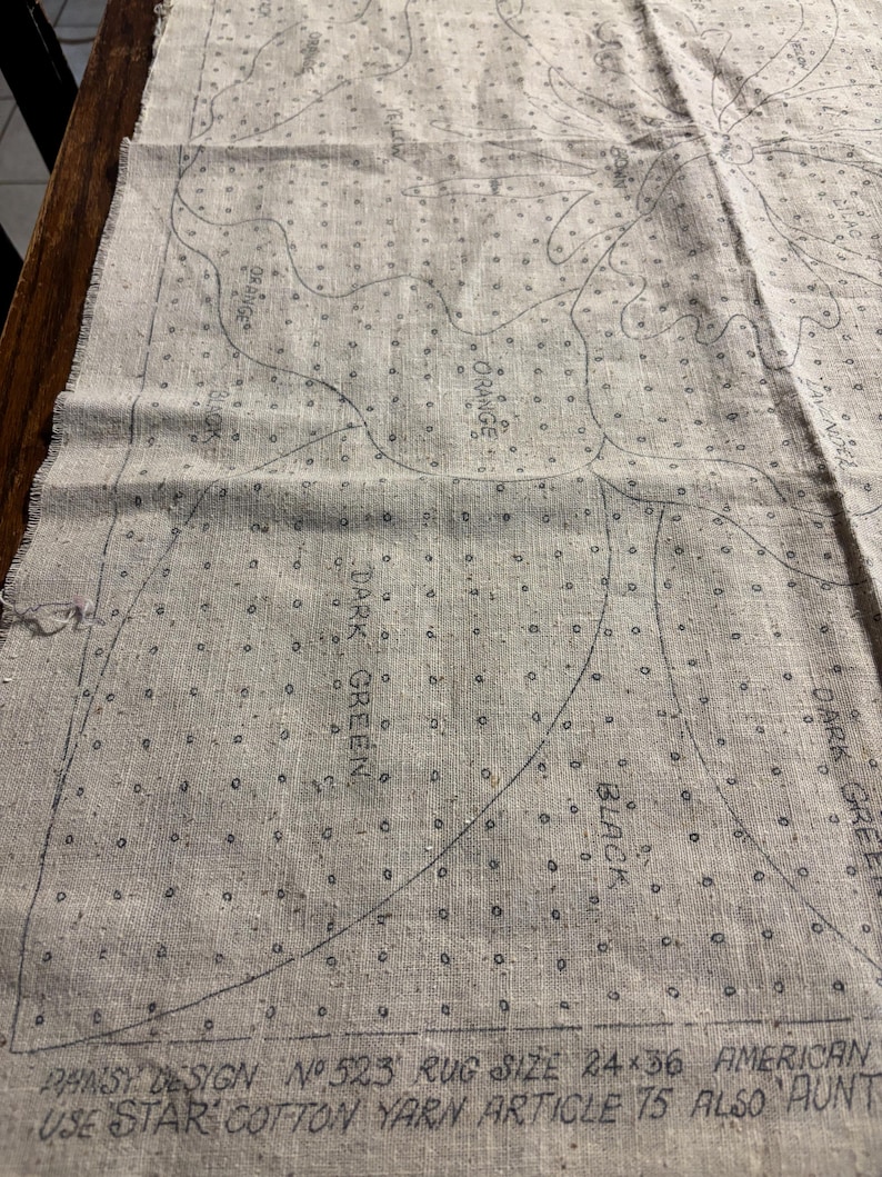 May include: A close-up of a beige canvas with a black and white design for a rug. The design includes a variety of shapes and lines, with the words "PANSY DESIGN N№ 523 RUG SIZE 24&times;36 AMERICAN USE STAR COTTON YARN ARTICLE 15 ALSO AUNT" printed at the bottom.