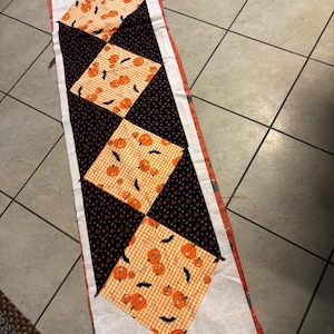 Op de afbeelding: Een Halloween-tafelloper met een patchwork-ontwerp. Het heeft afwisselende zwarte en oranje vierkanten met pompoen- en vleermuispatronen. De randen zijn afgewerkt met een witte rand en een oranje stof met patroon.