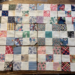 May include: A patchwork quilt top composed of alternating white squares and squares of patterned fabric. The fabric squares feature floral, plaid, and geometric designs in various colors, including blue, red, and green. The quilt top is laid out on a wooden surface.
