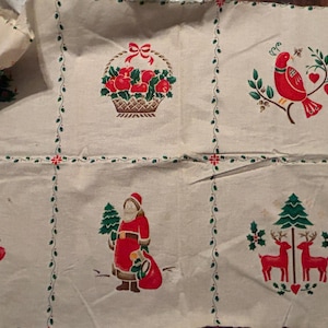 May include: Cream-colored fabric with embroidered Christmas-themed designs. Features a basket of red apples, a red bird on a branch, a goose with a wreath, Santa Claus, and two red deer with a Christmas tree.