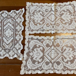 May include: Three antique, off-white lace doilies on a wooden surface. The doilies feature intricate floral and geometric patterns. One is rectangular, and two are square. The lace has a delicate, open weave, showcasing the detailed craftsmanship.