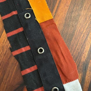 May include: Close-up of a patchwork textile with a variety of colors and textures. The design incorporates black fabric with red stripes, solid black fabric, orange, rust, and white. Metal eyelets are visible.