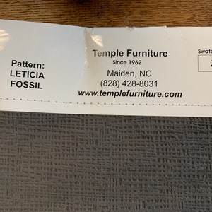 May include: A fabric swatch in a dark gray color with a textured woven pattern. The swatch is labeled "Leticia Fossil" and has a swatch number of 377. The label also includes the company name "Temple Furniture" and contact information.