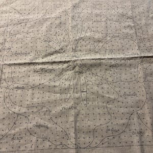 May include: A beige fabric with a black outline of a flower with the words "yellow", "orange", "lavender", "dark green", "black", "brown", and "lilac" written on it.