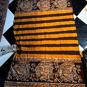 Op de afbeelding: Een rechthoekig textiel met een bruin en oranje gestreept patroon. De boven- en onderranden hebben een gedetailleerd ontwerp met vogelachtige figuren in wit en oranje. De stof ligt op een zwart-wit geblokte vloer.