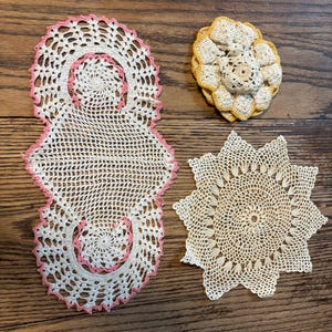 May include: Four handmade crochet doilies in various shapes and designs. One is a long, rectangular shape with pink trim, another is a flower shape with yellow trim, and two are circular with intricate patterns. The doilies are cream-colored.