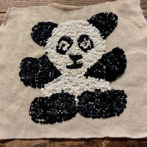 Könnte beinhalten: Ein beiger Stoffflicken mit einem Panda-Bär-Design aus Pailletten. Der Panda hat schwarze Paillettenohren, Pfoten und Umrisse, wobei weiße Pailletten das Gesicht und den Körper ausfüllen. Der Flicken ist rechteckig mit einem rohen Rand. Geeignet für Kleidung.