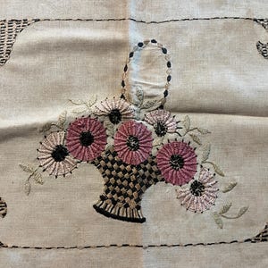 May include: A beige linen fabric with a black and brown embroidered design of a basket of pink and white flowers.
