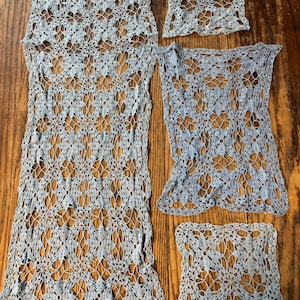 Peut inclure: Ensemble de pièces de dentelle crochetée bleu clair, comprenant un long chemin de table rectangulaire, deux pièces rectangulaires plus petites et deux pièces carrées. Le motif ajouré présente un motif floral. Les articles sont présentés sur une surface en bois.