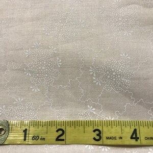 Pode incluir: Close-up de um tecido bege com um delicado padrão floral branco. Uma fita métrica amarela mostra 12,7 cm. O tecido tem uma textura subtil e é adequado para projetos de costura e artesanato.
