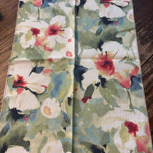 May include: A rectangular fabric swatch with a floral pattern. The design features large white flowers with red and pink centers, set against a backdrop of green, blue, and cream hues. The fabric has a textured appearance, and a label is attached.