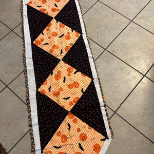 May include: A Halloween-themed table runner featuring alternating orange and black diamond patterns. The orange diamonds have a pumpkin and bat print, while the black diamonds have a small triangle pattern. The edges are trimmed with a patterned fabric.