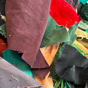 May include: A close-up of a pile of fabric scraps in various colours. The fabrics include corduroy and other textiles in shades of burgundy, green, red, black, and grey. The edges of the fabric are frayed.