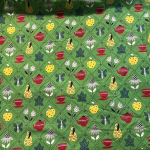 Op de afbeelding: Groene stof met een herhalend patroon van rode theepotten, kopjes, gele appels, peren, vlinders en witte bloemen. Het ontwerp is geplaatst op een raster van vorken en ruiten.